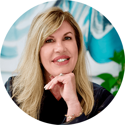 A woman with long blonde hair smiling, wearing a black jacket, and resting her chin on her hand. The background features abstract blue and white artwork and a green plant.